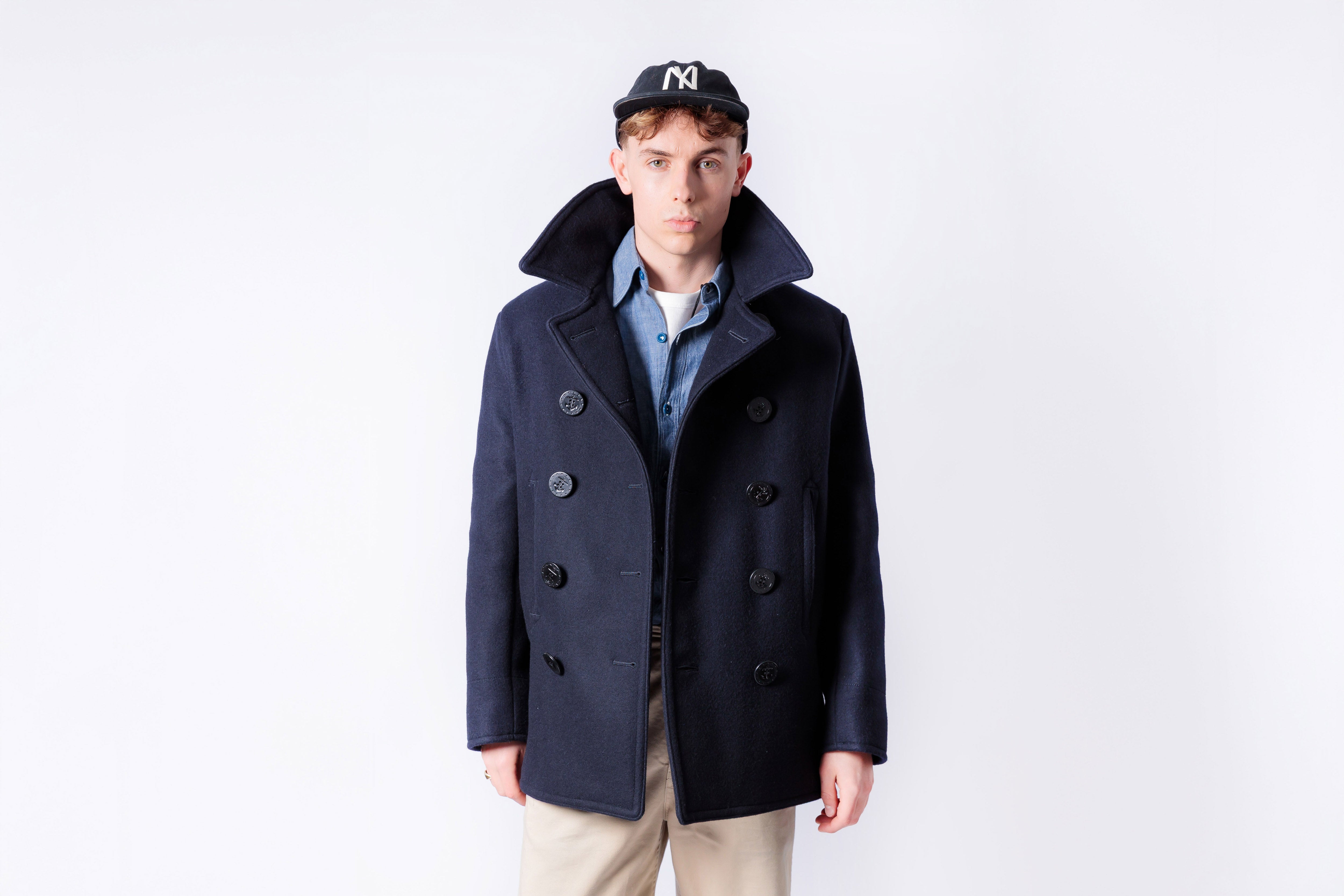 Man wearing a navy peacoat and cap on a white background