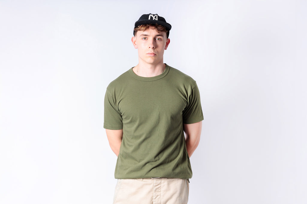 Person wearing an olive green t-shirt and beige pants against a white background