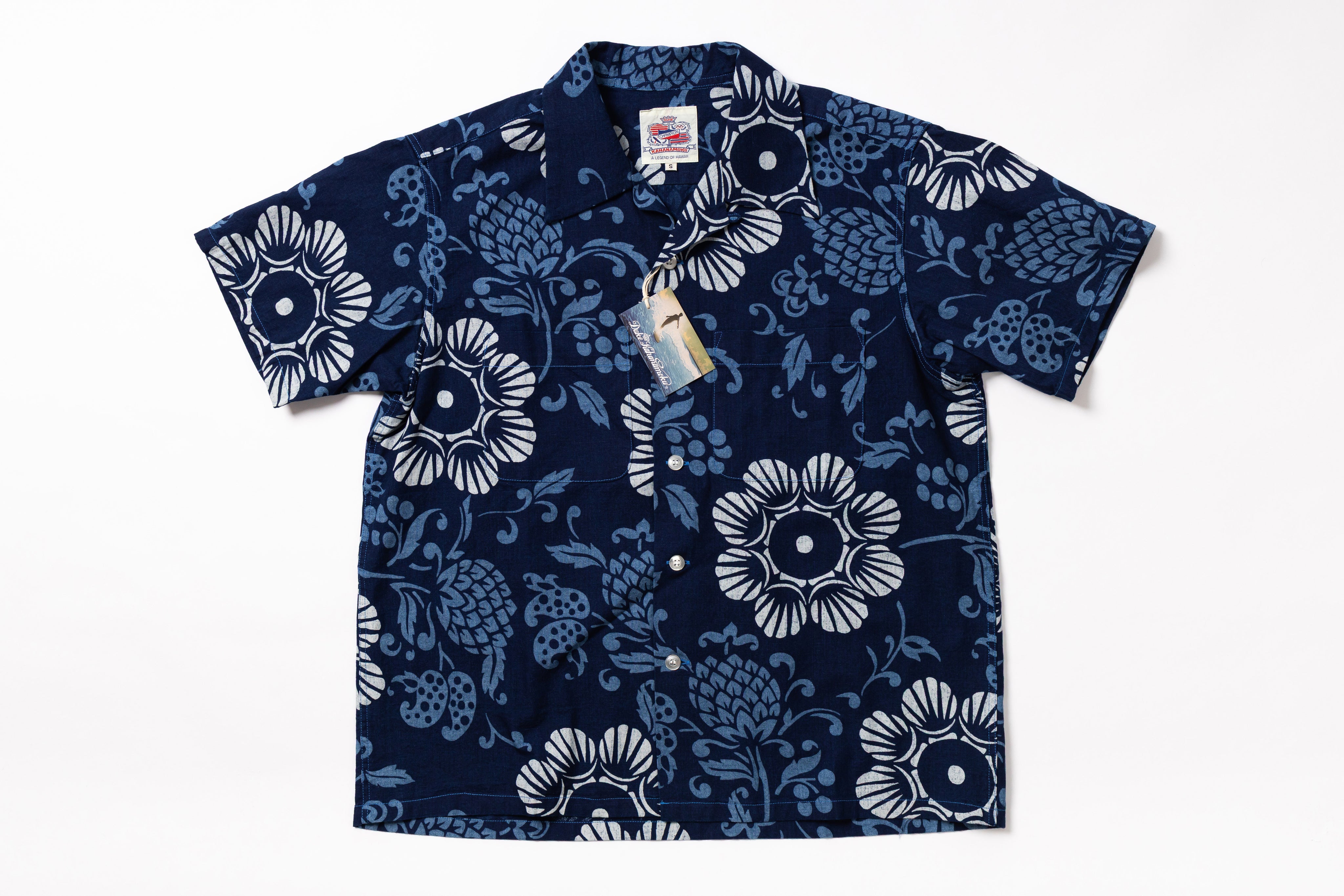 A short-sleeve button-up camp shirt in navy blue with a white floral and pinecone pattern, shown flat against a white background with retail tags attached.