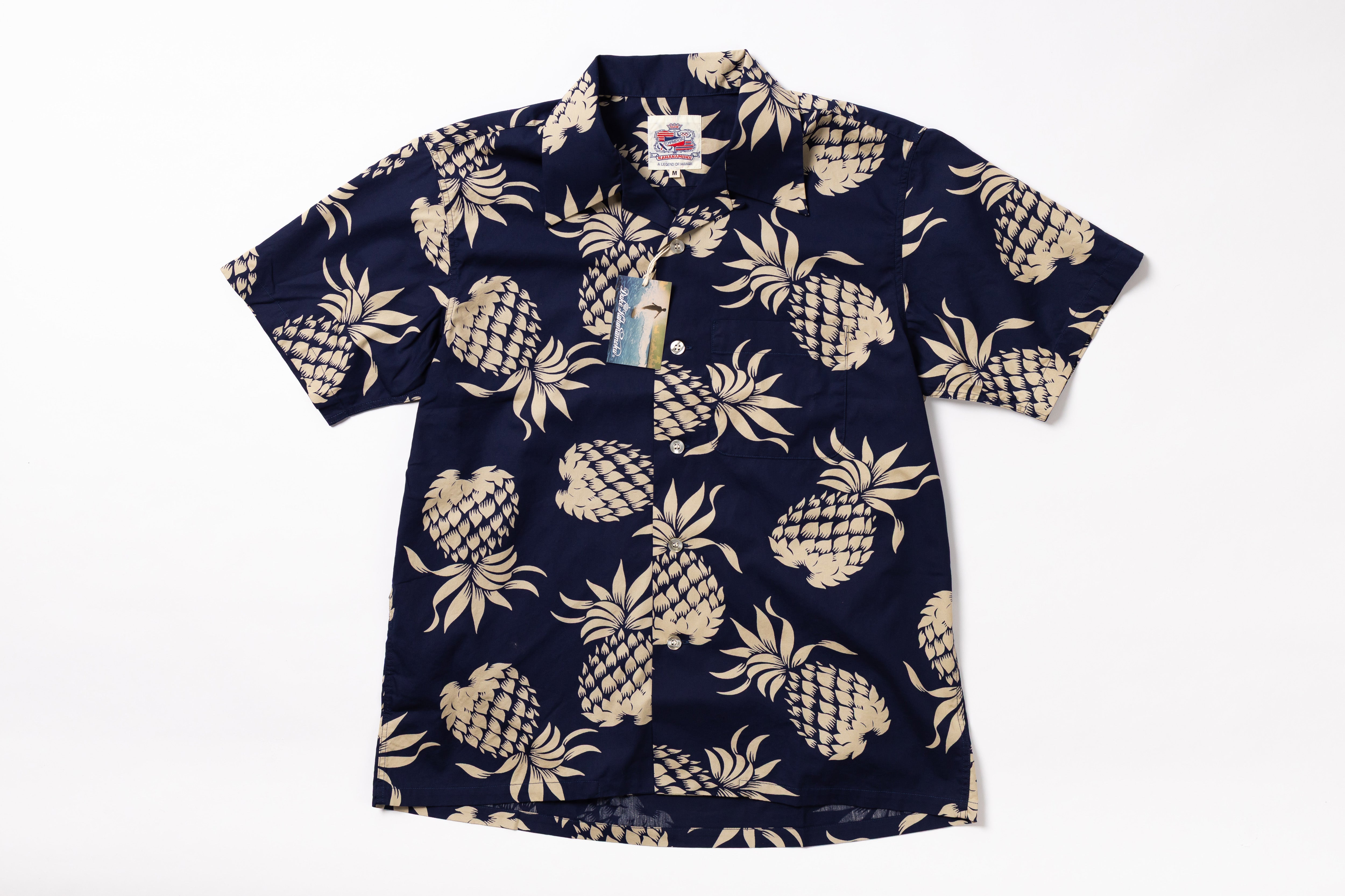 A short-sleeve button-up camp shirt with a navy blue base and a beige pineapple pattern, shown flat against a white background with a hangtag attached.