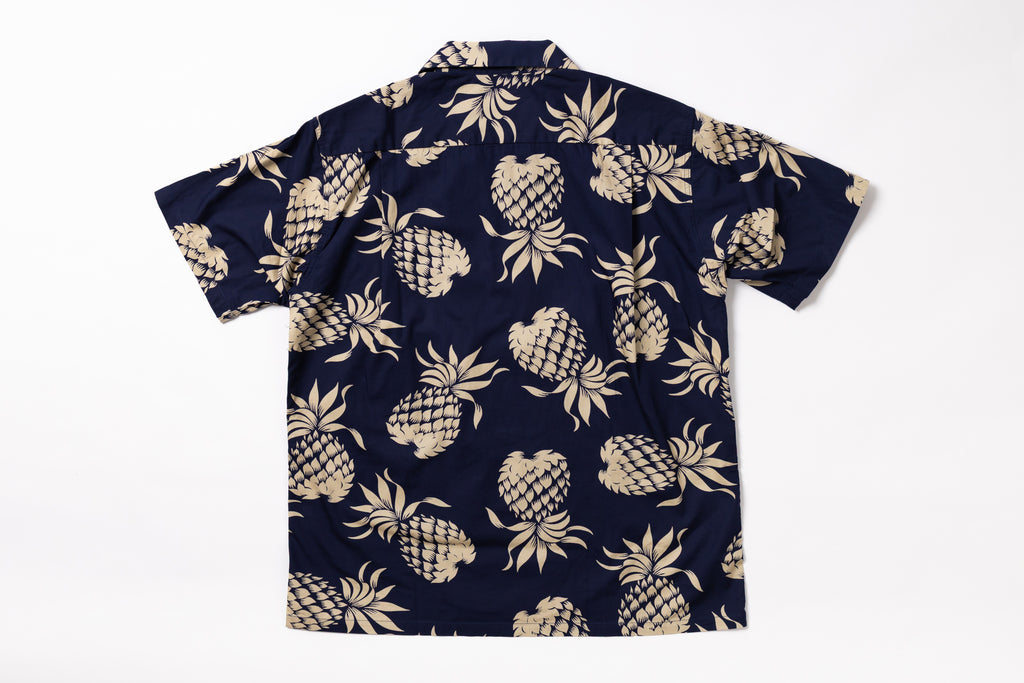 A short-sleeve button-up shirt with an all-over beige pineapple print on a dark navy blue background, laid flat against a white background.