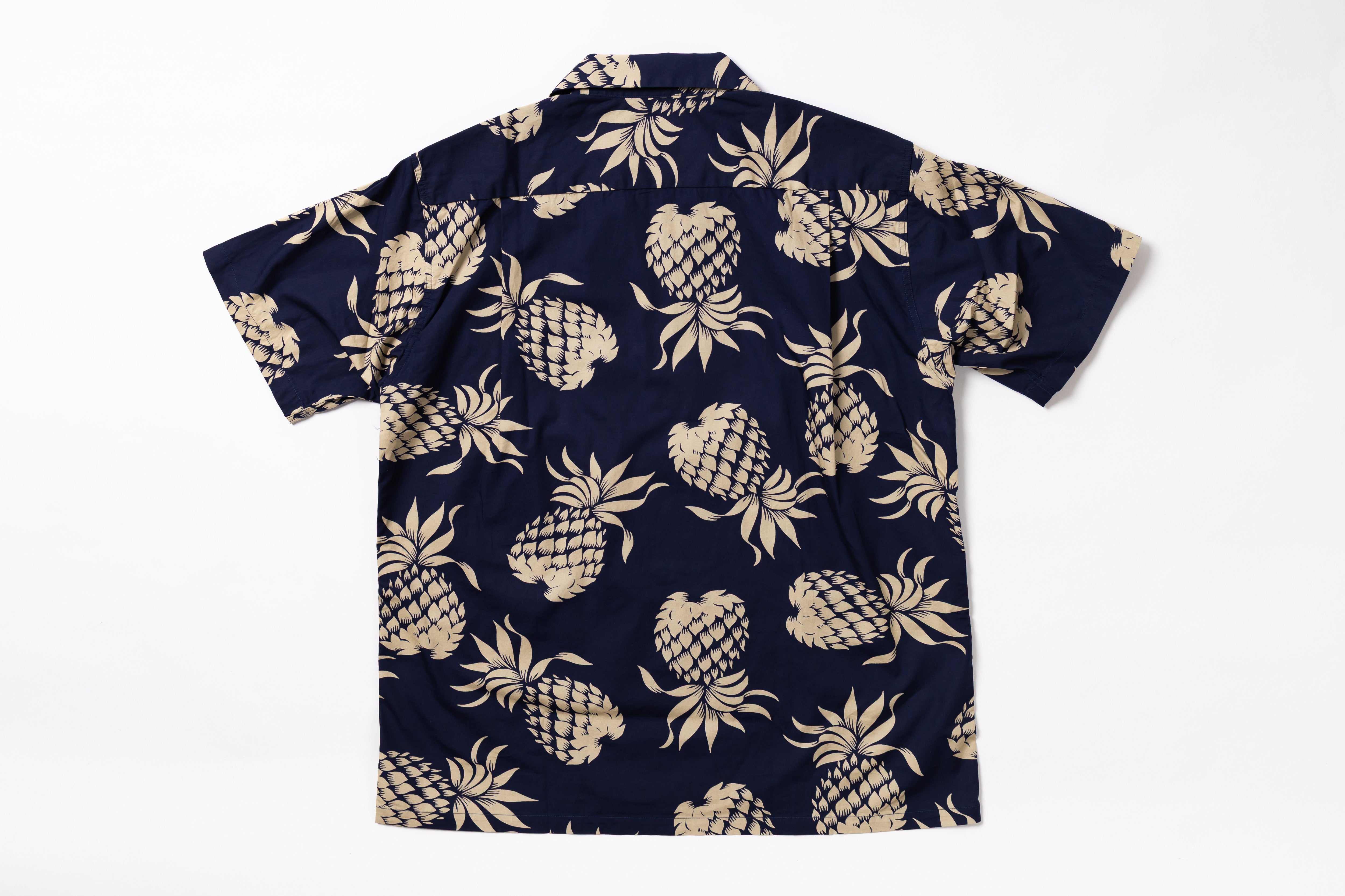 A short-sleeve button-up shirt with an all-over beige pineapple print on a dark navy blue background, laid flat against a white background.