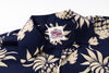 Close-up of a navy blue short-sleeve button-up shirt with a beige tropical floral print, featuring a visible brand label on the collar.