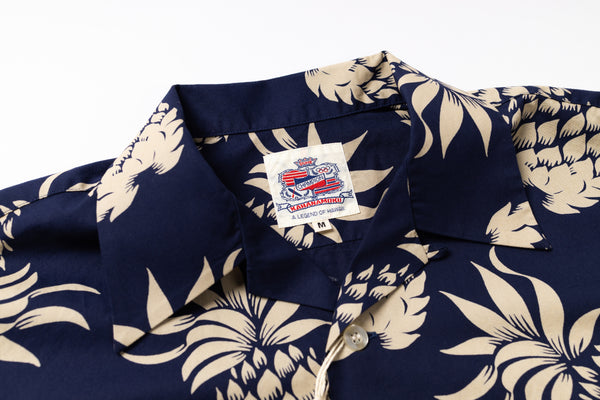 Close-up of a navy blue short-sleeve button-up shirt with a beige tropical floral print, featuring a visible brand label on the collar.