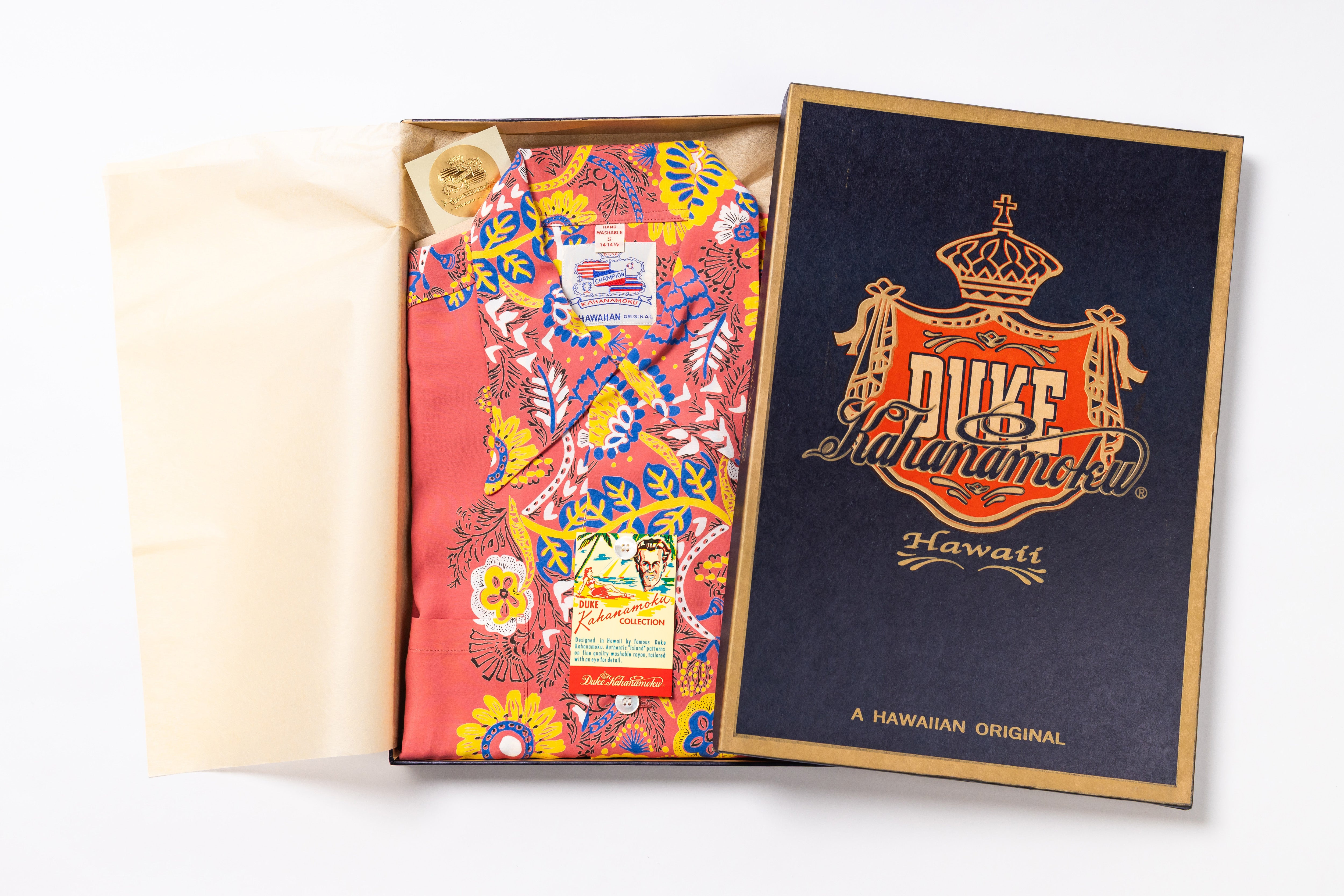 An open presentation box showing a folded Hawaiian shirt with a bright multicolor floral and paisley print, alongside the branded navy and gold Duke Ahuanahe Hawaii box lid on a white background.