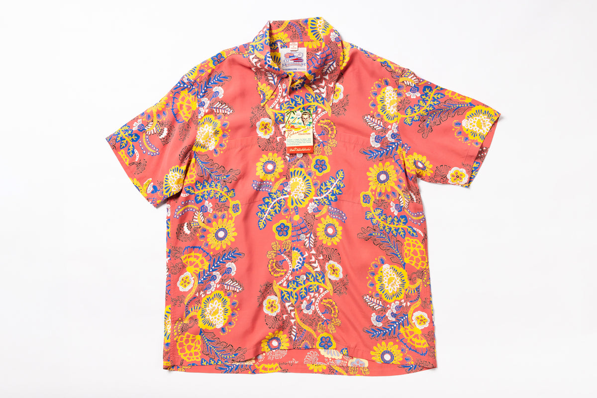 A short-sleeve button-up camp shirt in a coral pink base with a vibrant multicolor paisley and floral print in yellow, blue, and orange, laid flat against a white background.