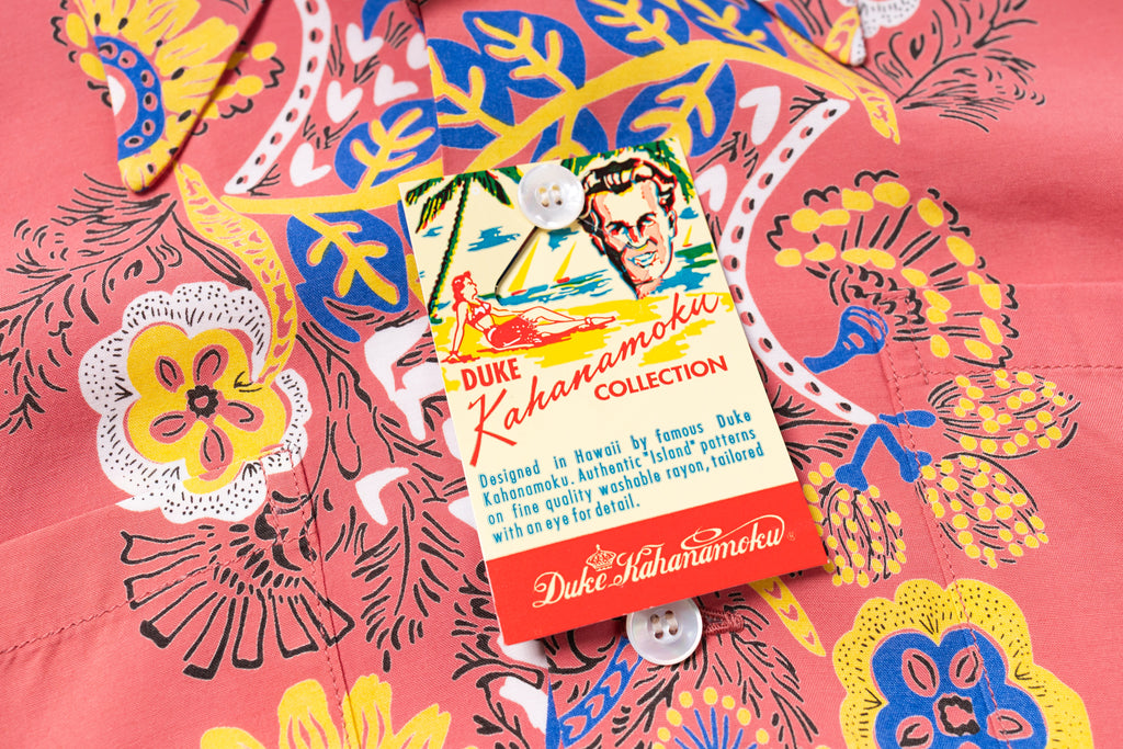 A close-up of a coral pink Hawaiian shirt with a bold multicolor floral and bird print, featuring a branded hangtag for Duke Kahanamoku Collection.