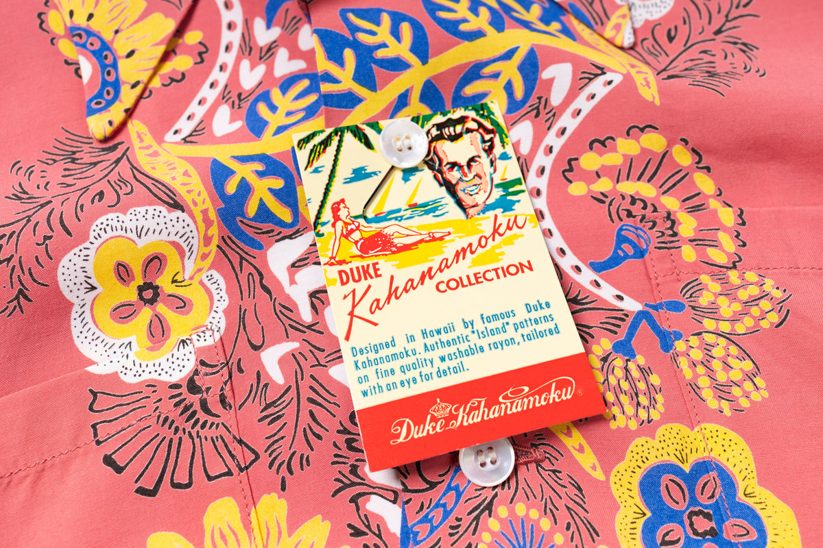A close-up of a coral pink Hawaiian shirt with a bold multicolor floral and bird print, featuring a branded hangtag for Duke Kahanamoku Collection.