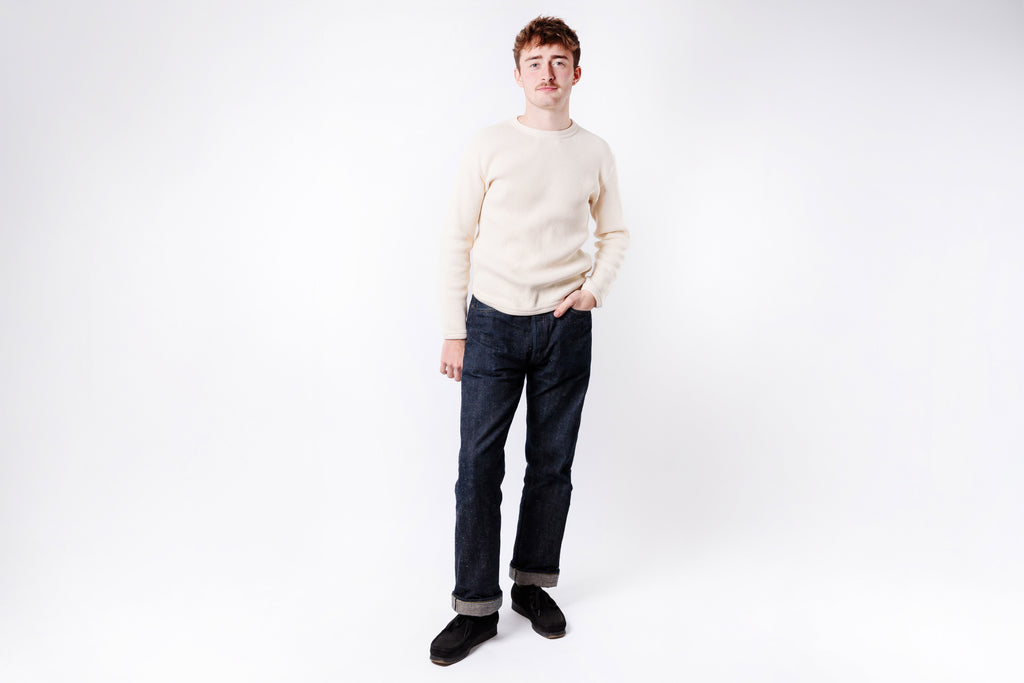 Man wearing a beige sweater and dark jeans on a white background