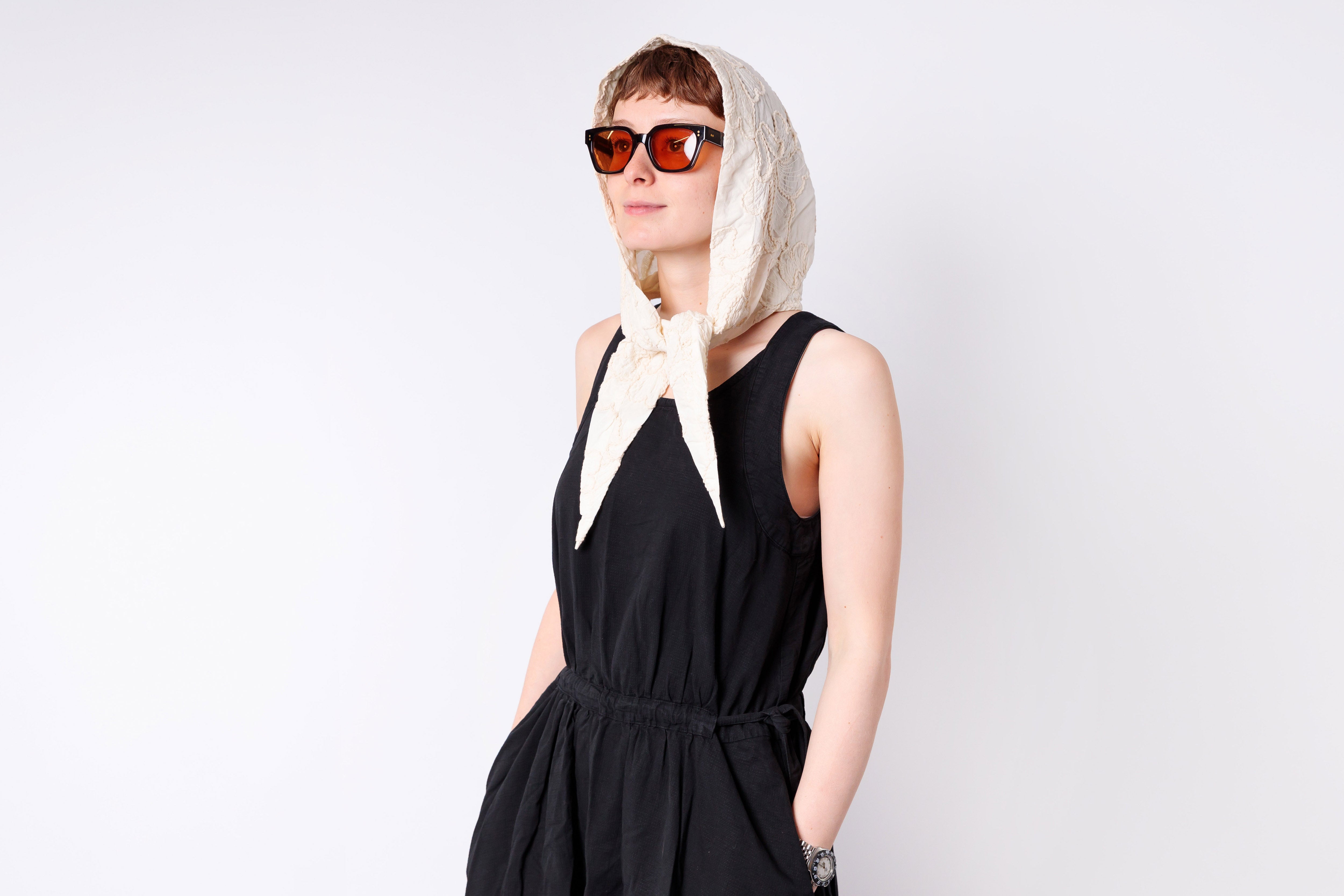Woman wearing a black dress, sunglasses, and a white scarf on a plain background