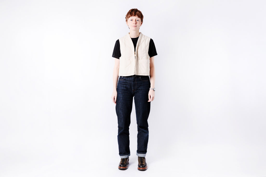 Person wearing a black shirt, beige vest, dark jeans, and brown shoes on a white background