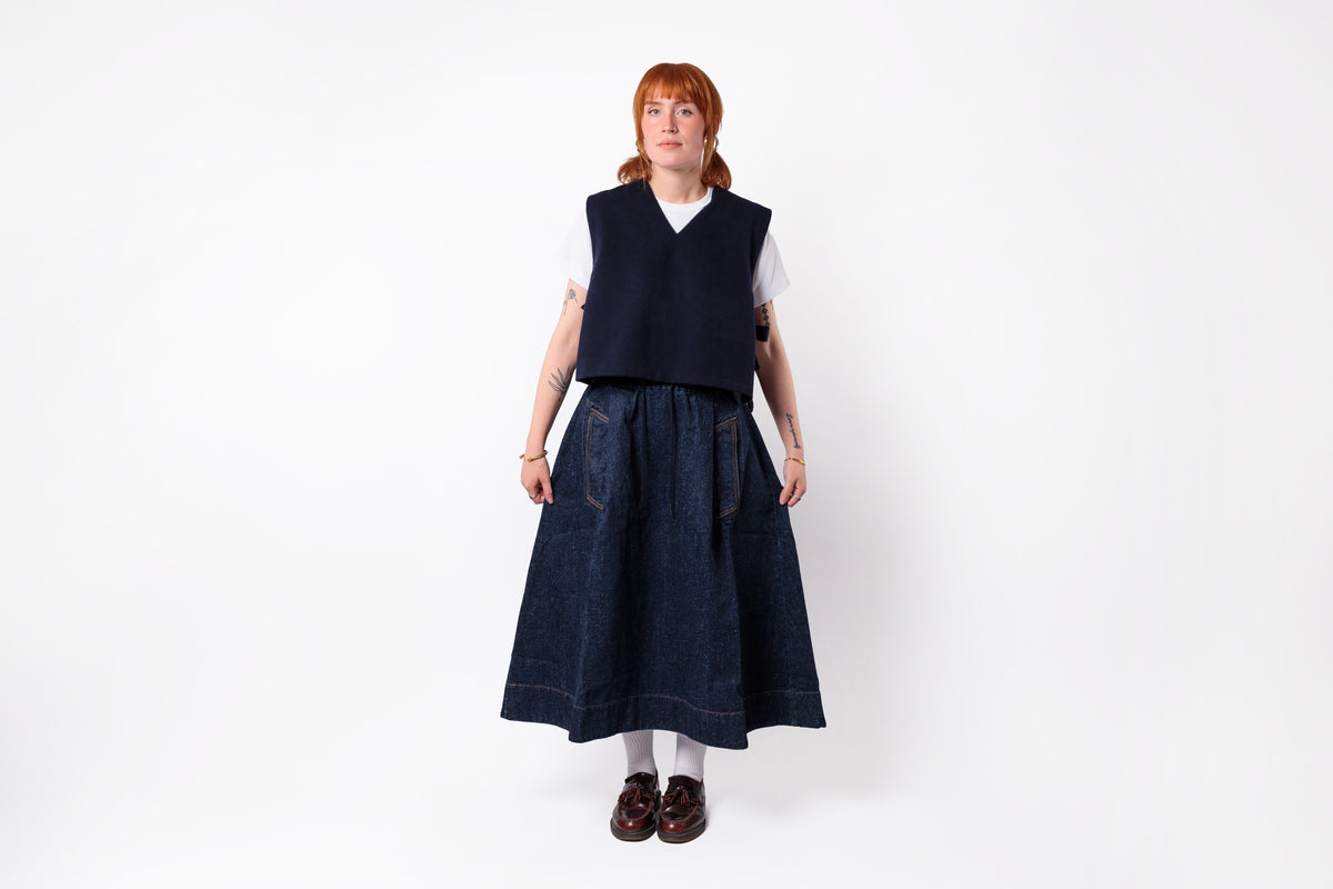 A model stands against a plain white studio background wearing a dark navy sleeveless vest over a white short-sleeve top, paired with a dark blue denim skirt, white tights, and dark brown shoes.