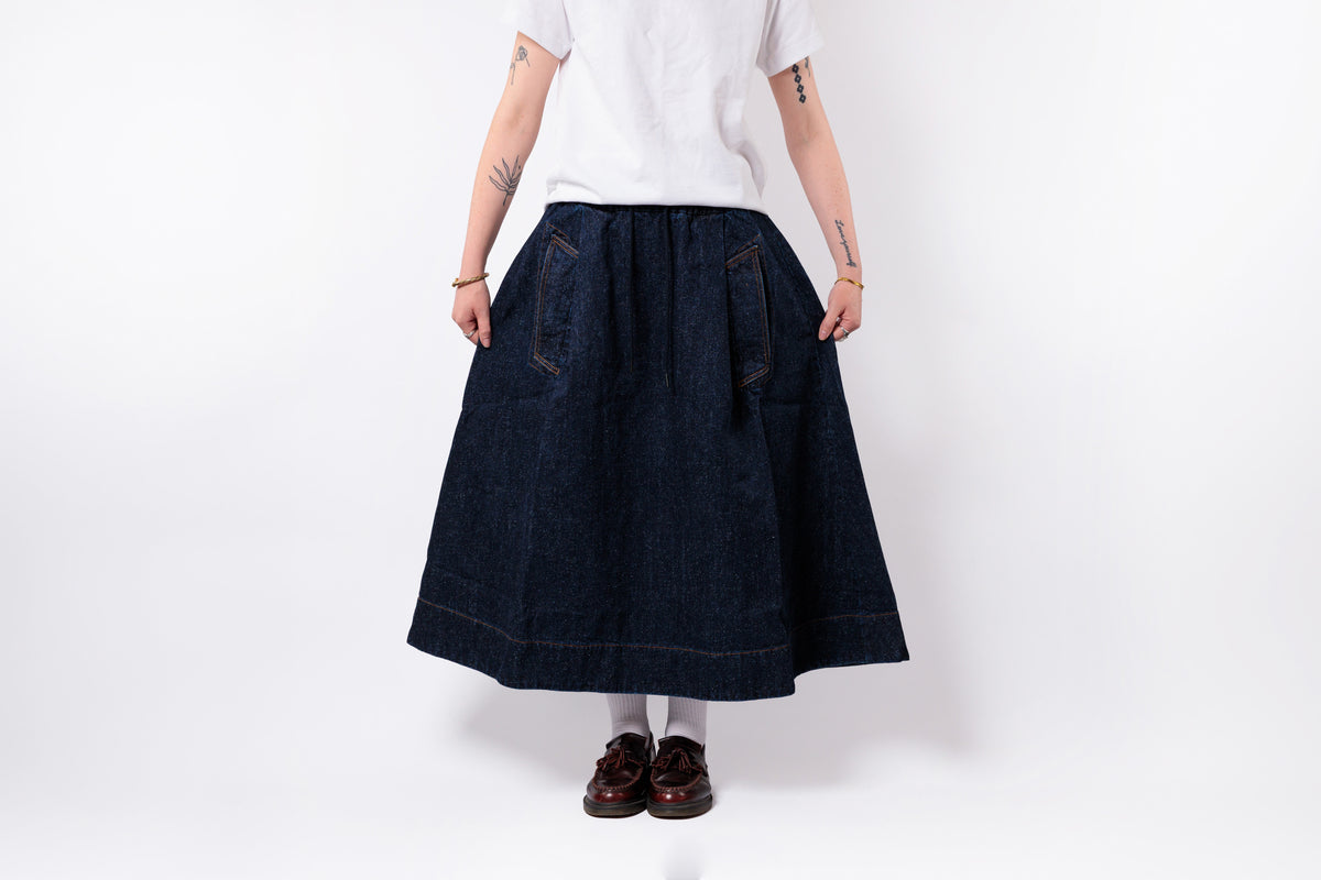 A person shown from the back wearing a dark indigo denim midi skirt with front patch pockets, paired with a white t-shirt, white socks, and dark brown loafers, against a plain white studio background.