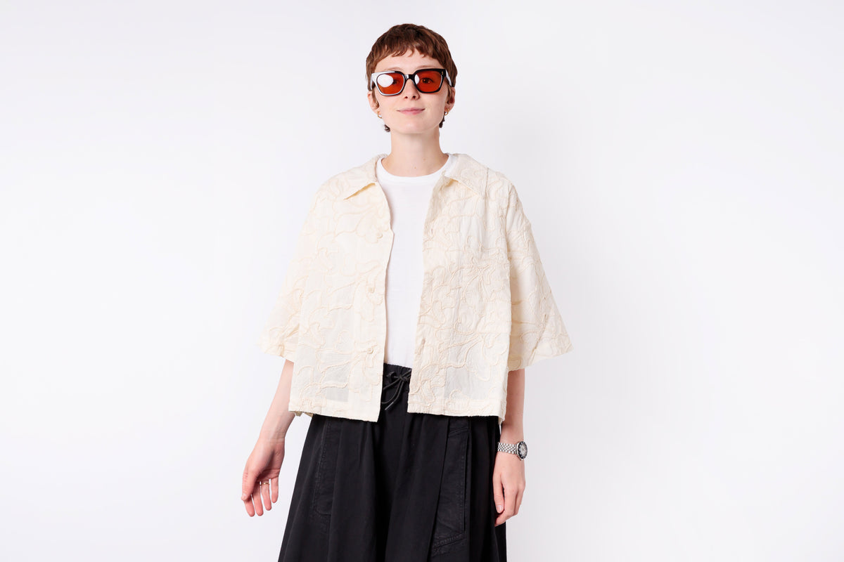Person wearing a cream jacket over a white shirt with black pants on a white background