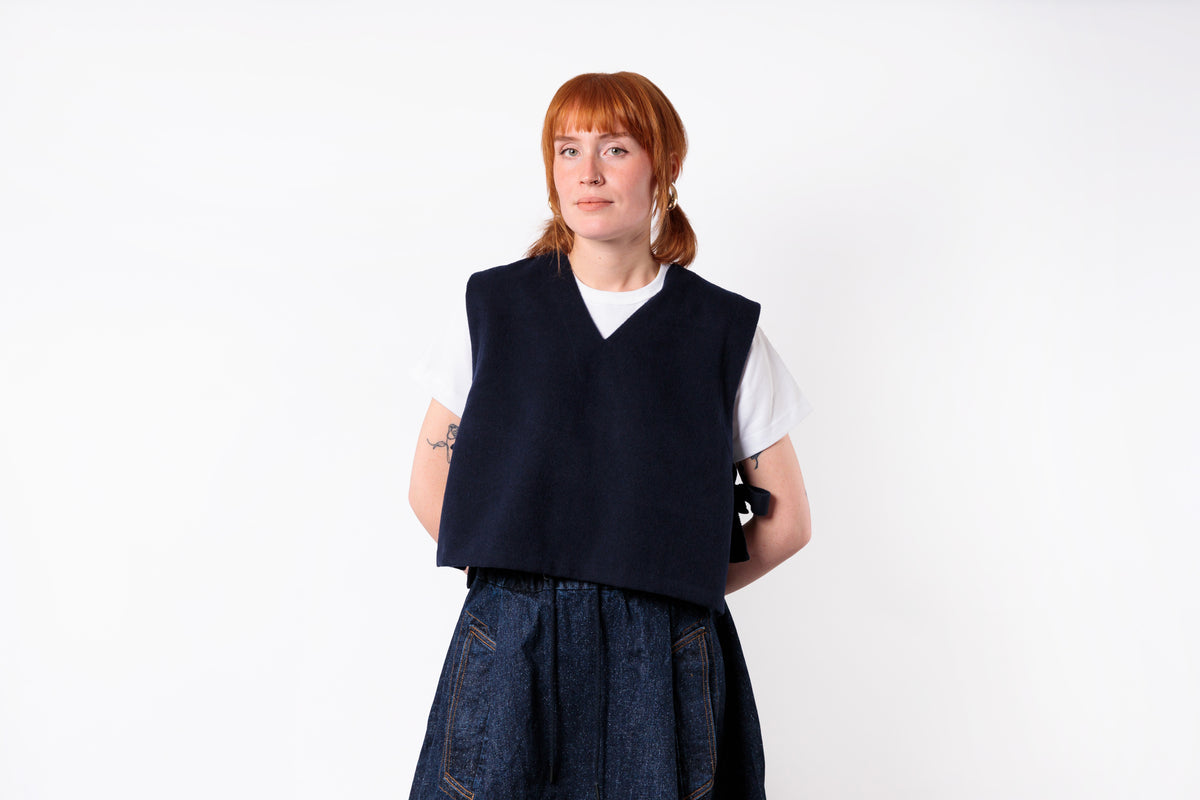 A woman with red hair wearing a dark navy sleeveless vest over a white t-shirt and a dark blue denim skirt, standing against a plain white background.