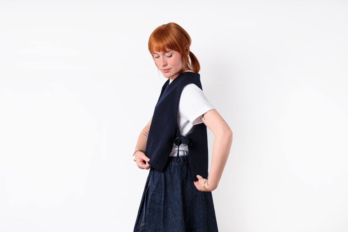 A woman with red hair tied back wears a dark navy sleeveless vest over a white short-sleeve top and a dark blue skirt, posing against a solid white studio background.