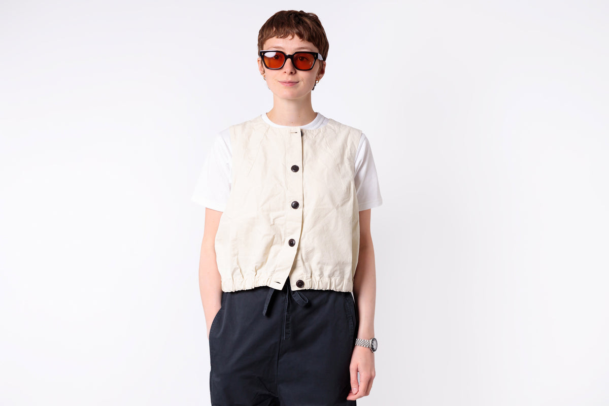 Person wearing a cream vest over a white shirt with dark pants on a white background