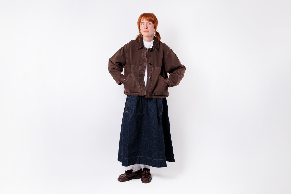 A model with red hair stands against a white studio background wearing a brown corduroy jacket over a white top, paired with a dark blue denim skirt and brown shoes.