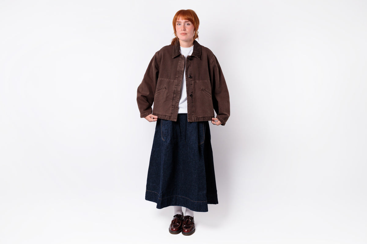 A person stands against a plain light gray studio background wearing a dark brown jacket over a white top, a long dark navy skirt, and dark brown shoes.