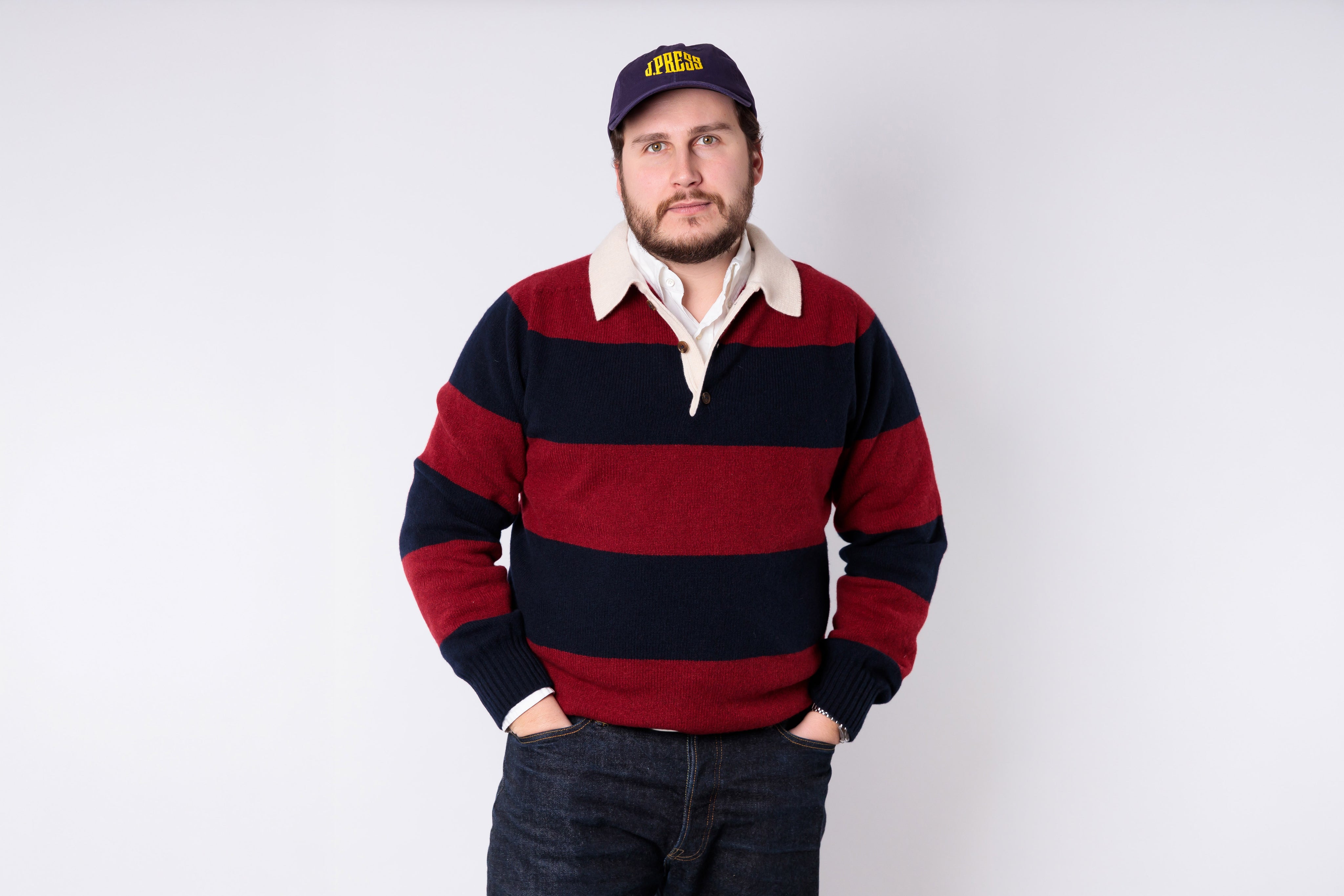 Man wearing a red and navy striped sweater with a white shirt underneath, standing against a plain background.