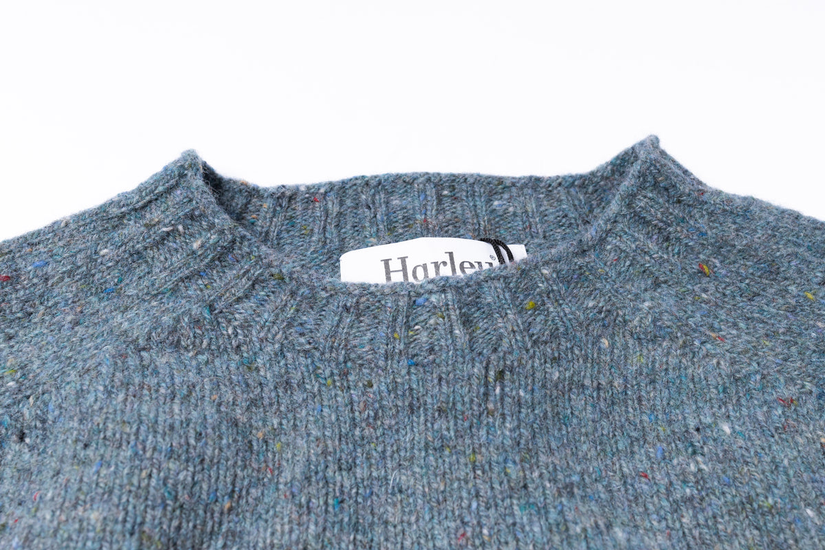 Close-up of a textured knit sweater in a heathered blue-gray color with flecks of multicolored yarn, featuring a white neck label with the brand name Harley.