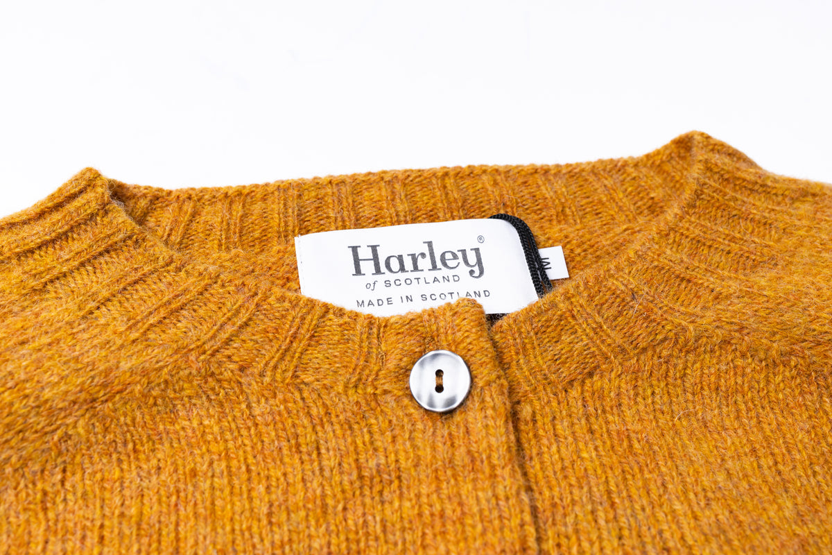 Close-up of a knit cardigan sweater in a heathered mustard yellow-orange color, featuring a white brand label that reads 'Harley of Scotland Made in Scotland' and a single silver button at the neckline.