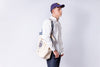 Person wearing a white shirt, blue jeans, and a purple cap with a beige backpack on a light gray background