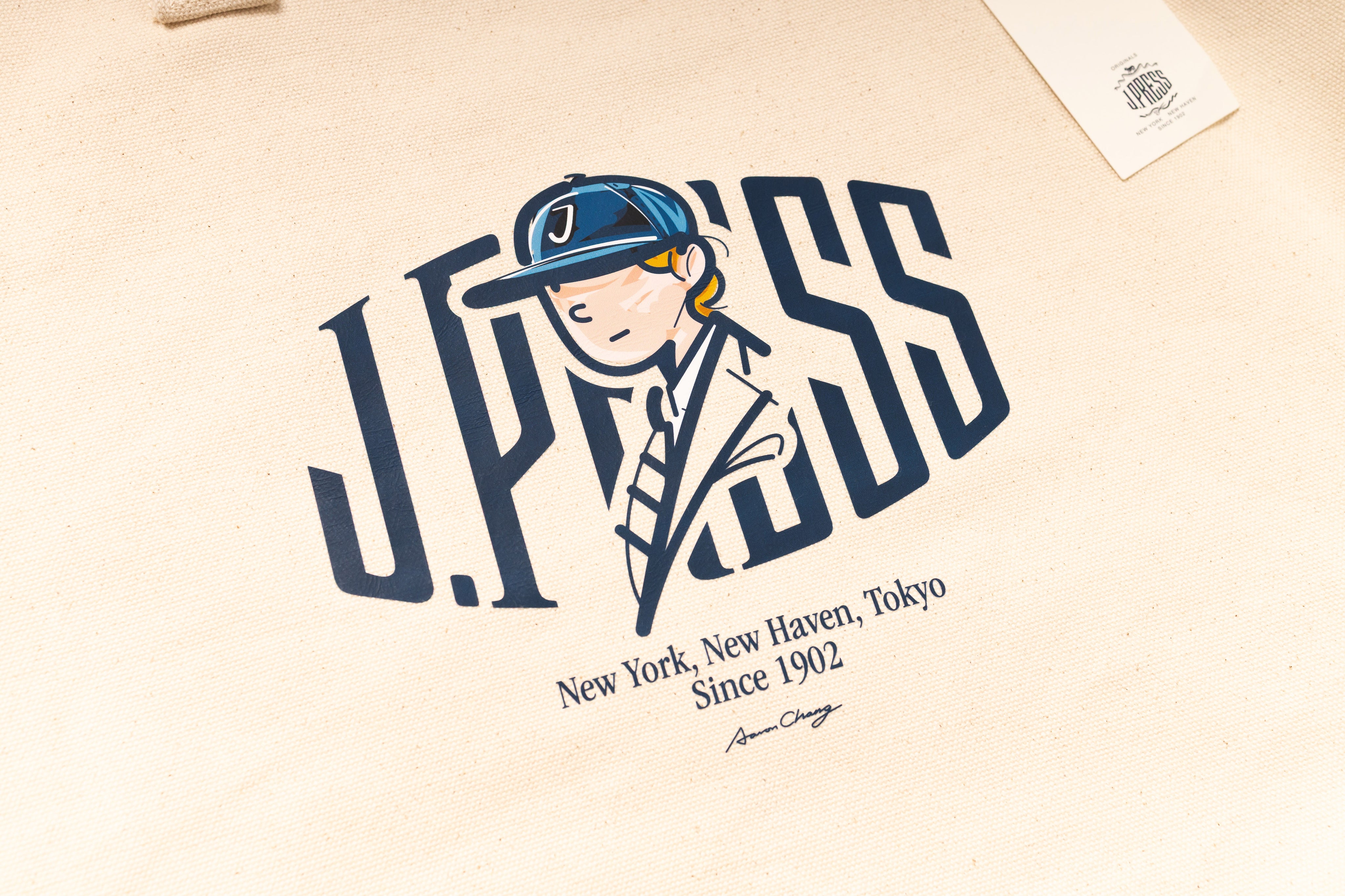 Beige tote bag with a graphic of a person wearing a cap and the text 'J.PRESS' on a beige background.