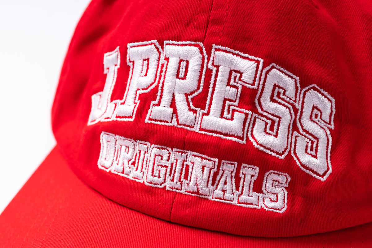 A close-up of a red baseball cap featuring white embroidered collegiate-style lettering that reads 'J.PRESS ORIGINALS' on the front panel.