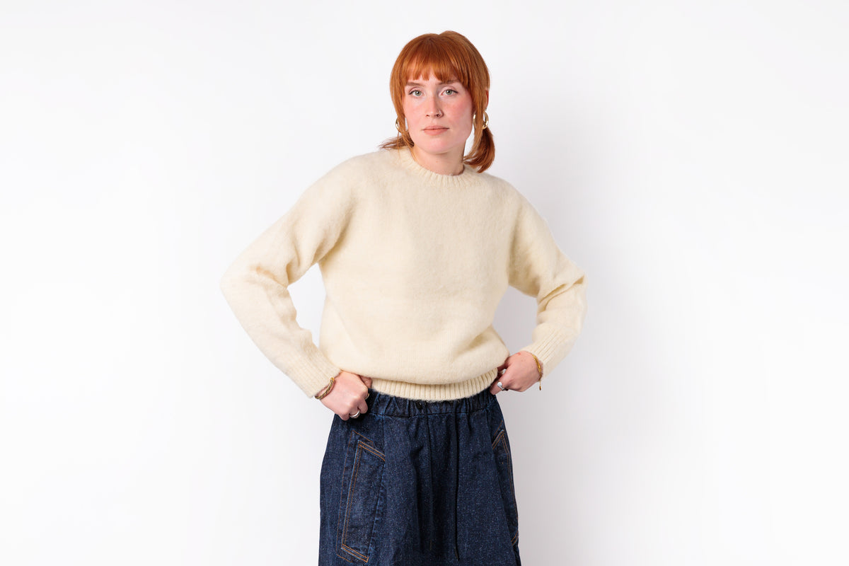 A woman with red hair wearing a cream-colored knit sweater and dark blue jeans, standing against a plain white background.