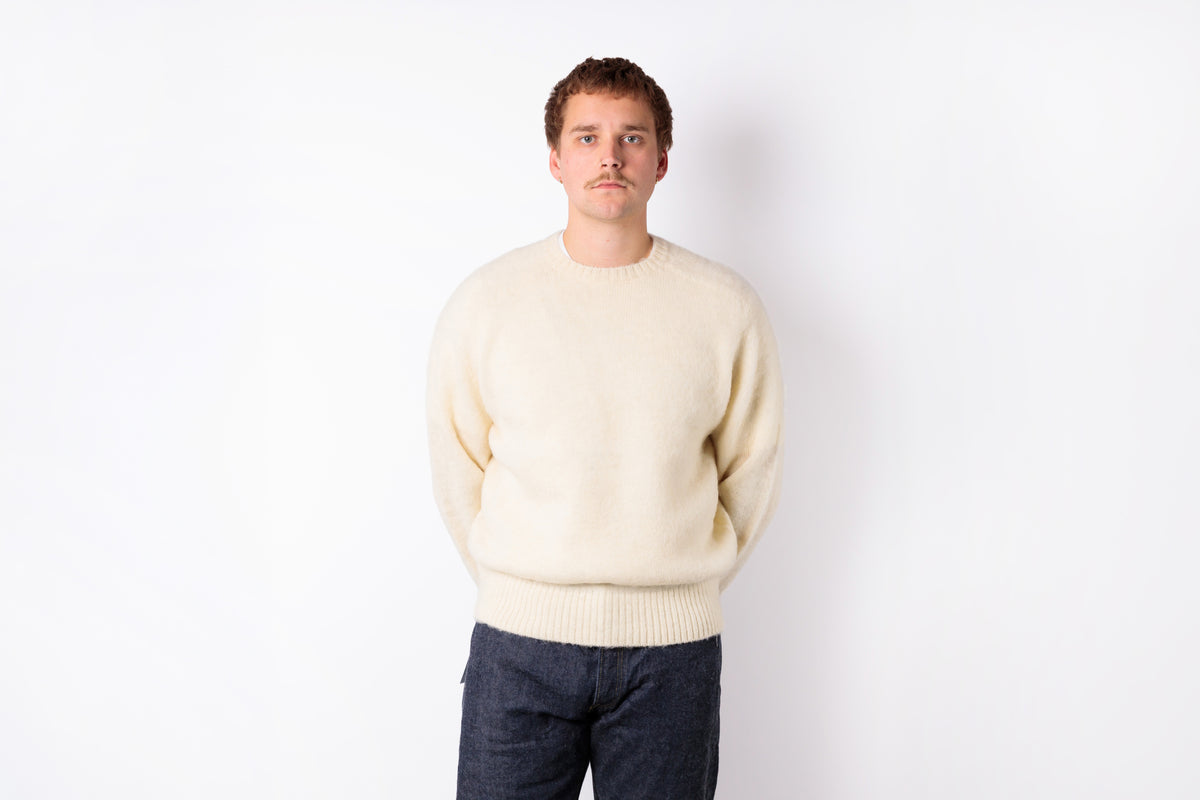 A man wearing a cream-colored knit crewneck sweater and dark blue jeans, standing against a plain white background.