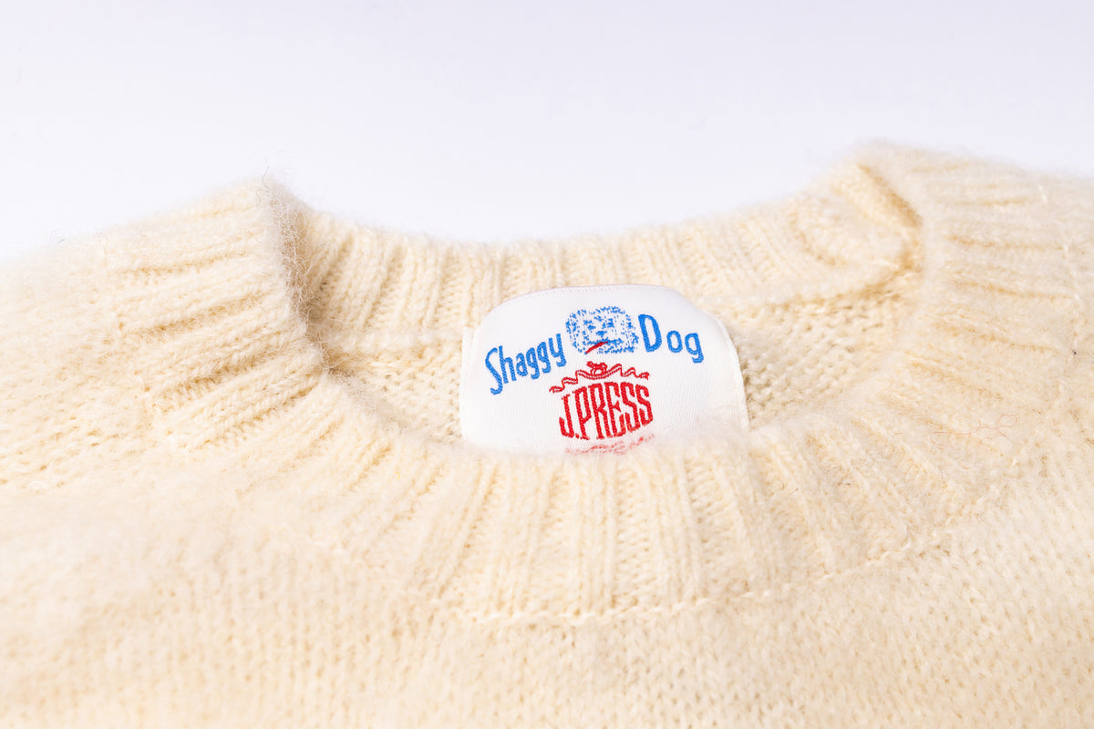 Close-up of a cream-colored knit sweater with a visible white neck label reading 'Shaggy Dog Press'.
