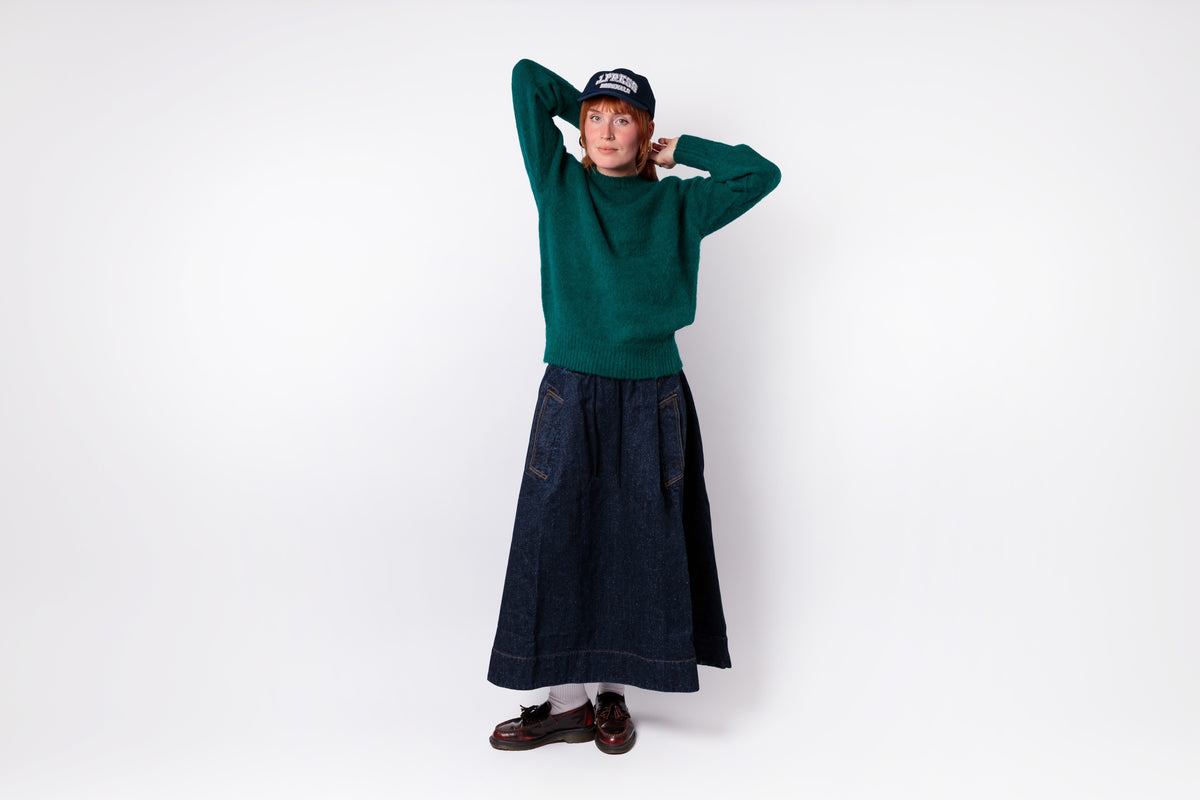 A person modeling a dark green long-sleeve knit sweater and a long dark blue denim skirt, paired with a black baseball cap, white socks, and brown loafers, against a plain white studio background.