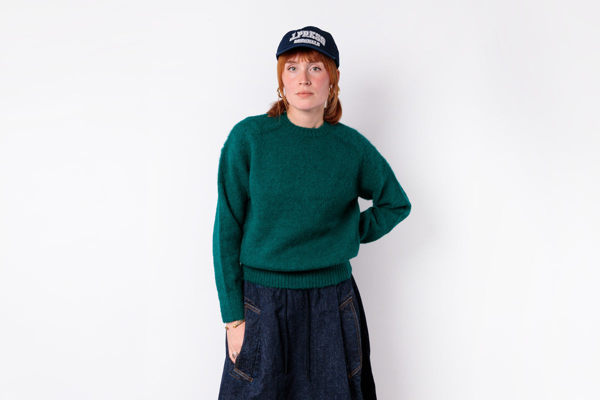 A woman wearing a dark green knit sweater, dark blue jeans, and a navy blue baseball cap with white text, standing against a plain white studio background.