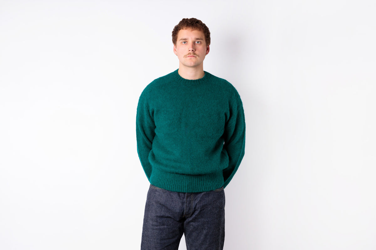 A man wearing a dark green knit crewneck sweater and dark gray jeans, standing against a plain white background.