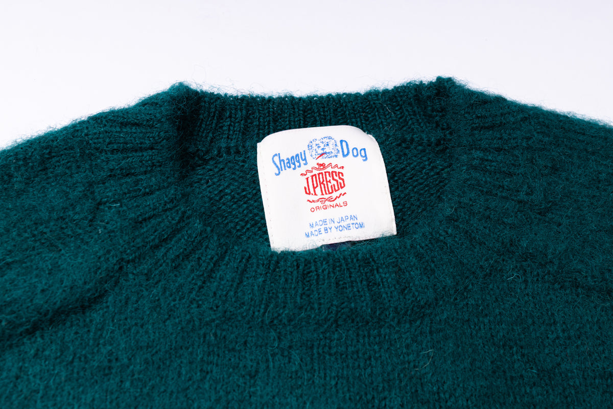 Close-up of a dark teal knit sweater showing the inner collar label with 'Shaggy Dog' and branding on a white background.