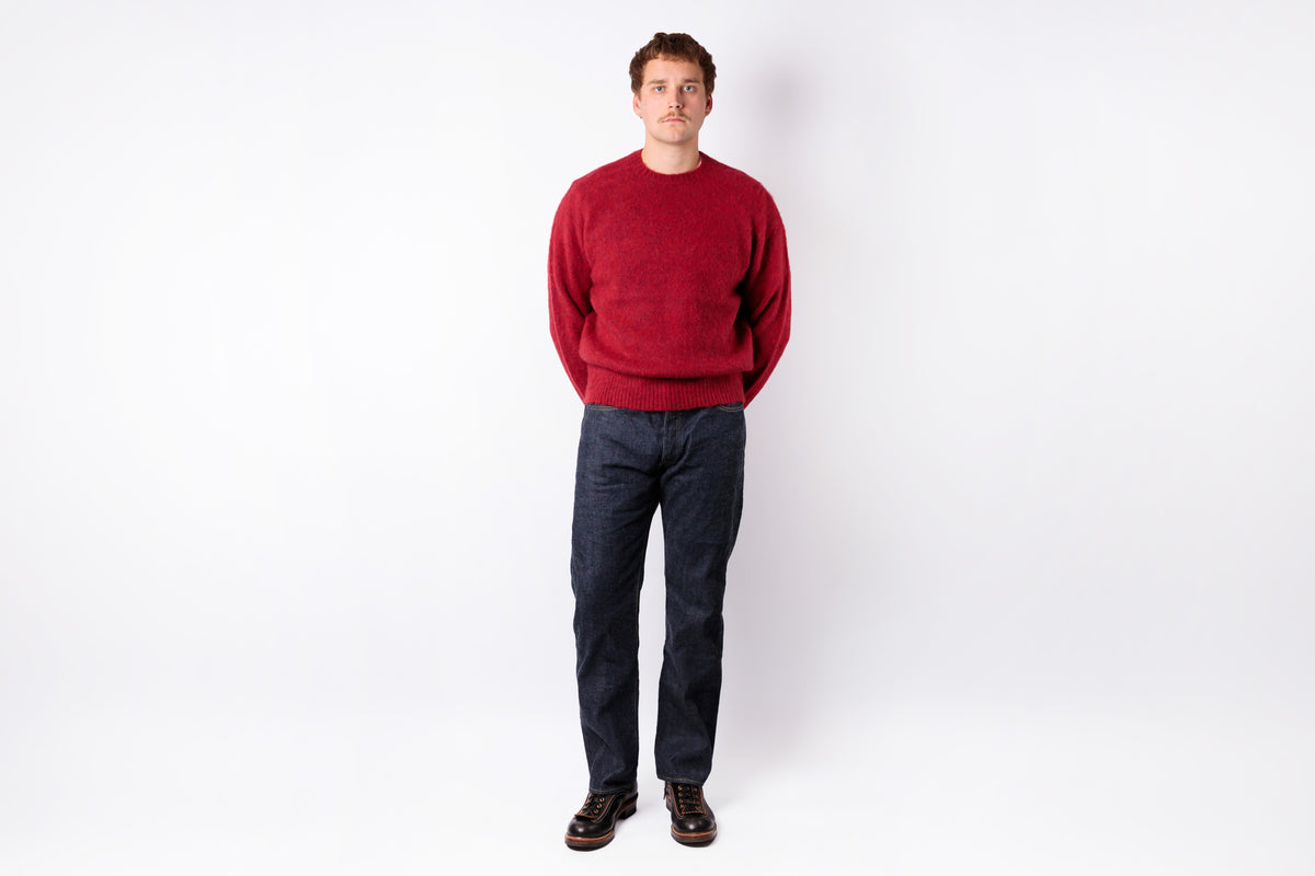 A man wearing a red crewneck sweater, dark blue jeans, and brown leather shoes, standing against a plain white studio background.
