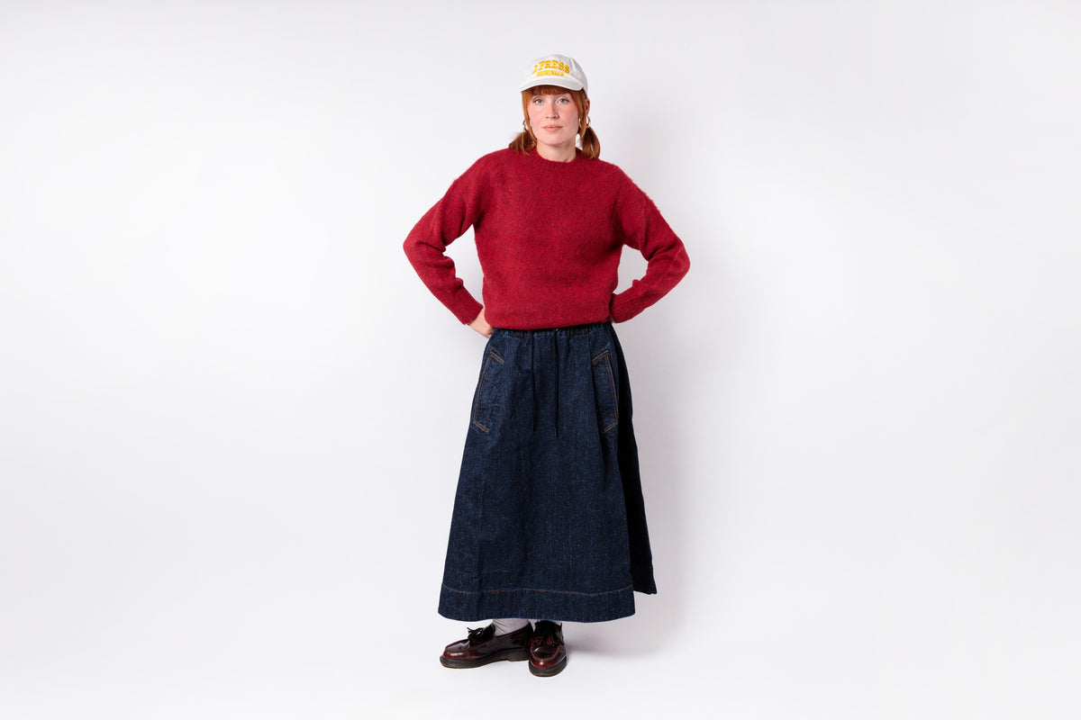 A person stands against a plain light gray studio background wearing a red knit sweater, a long dark denim skirt, dark brown shoes, and a white cap with yellow text.