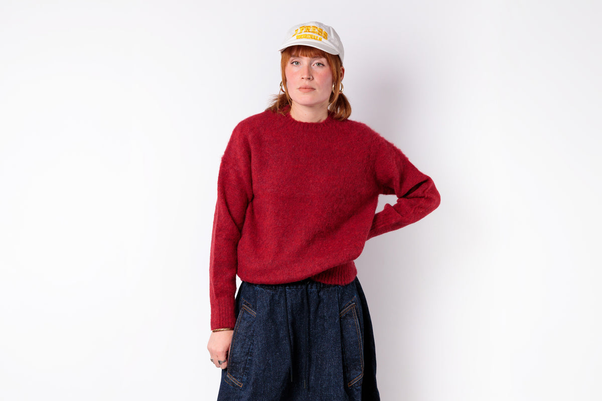 A woman modeling a fuzzy, textured red crewneck sweater, paired with dark blue jeans and a beige baseball cap with yellow lettering, set against a plain white studio background.