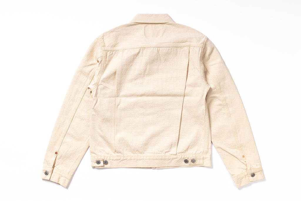 A light beige canvas utility jacket shown from the back, featuring a collar, long sleeves, and button details on the cuffs and hem, laid flat against a white background.