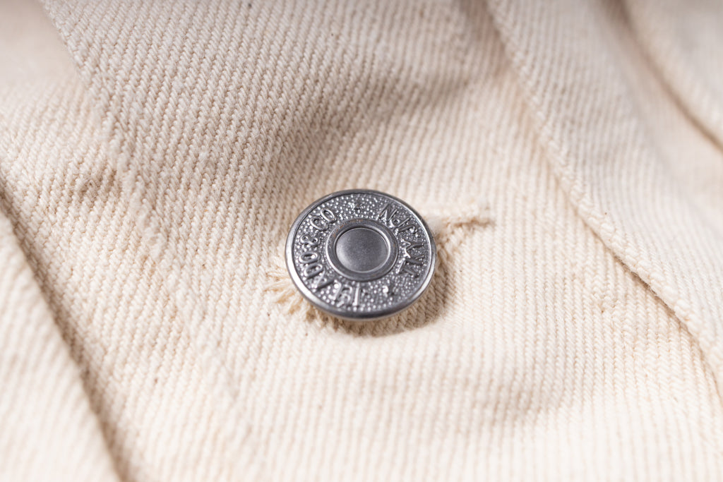 A close-up shot of a silver-toned metal snap button fastening a light beige or cream-colored canvas jacket.