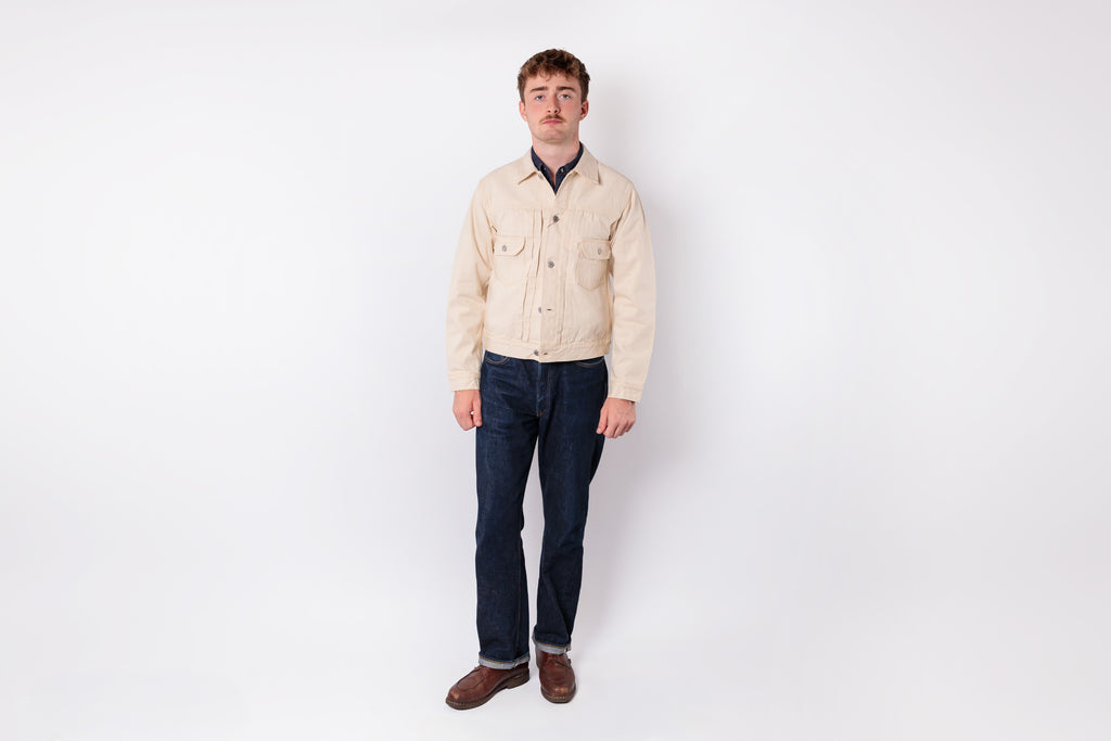 A man stands against a plain white studio background wearing a light beige button-up overshirt over a dark navy collared shirt, dark blue jeans, and brown shoes.