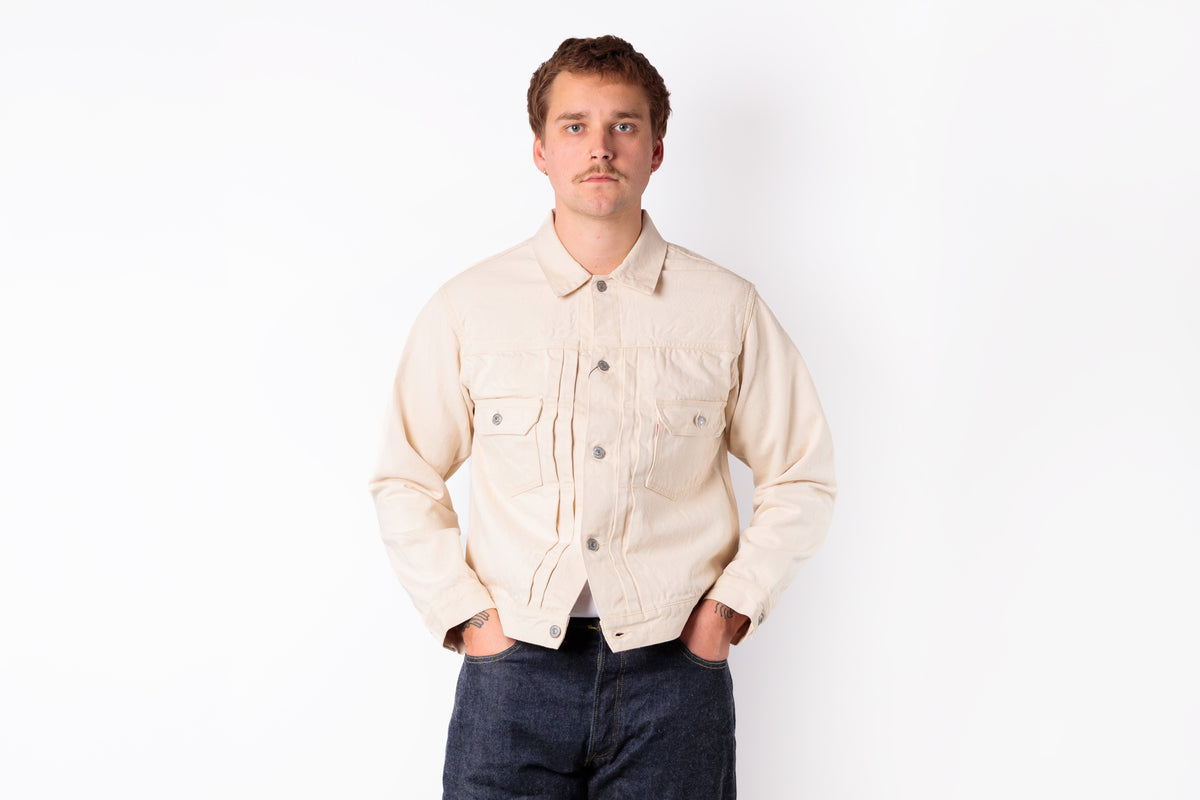 A man wearing a light beige denim trucker jacket and dark blue jeans, standing against a plain white background.