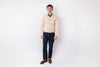A man stands against a plain white studio background wearing a light beige button-up overshirt over a dark navy collared shirt, dark blue jeans, and brown shoes.