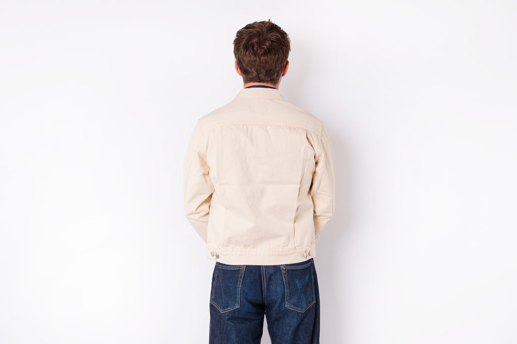 A man viewed from behind wearing a light beige canvas utility jacket and dark blue jeans, standing against a plain white background.