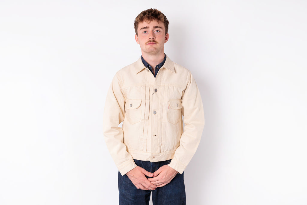 A man wearing a light beige canvas utility jacket over a dark navy collared shirt and dark blue jeans, standing against a plain white studio background.