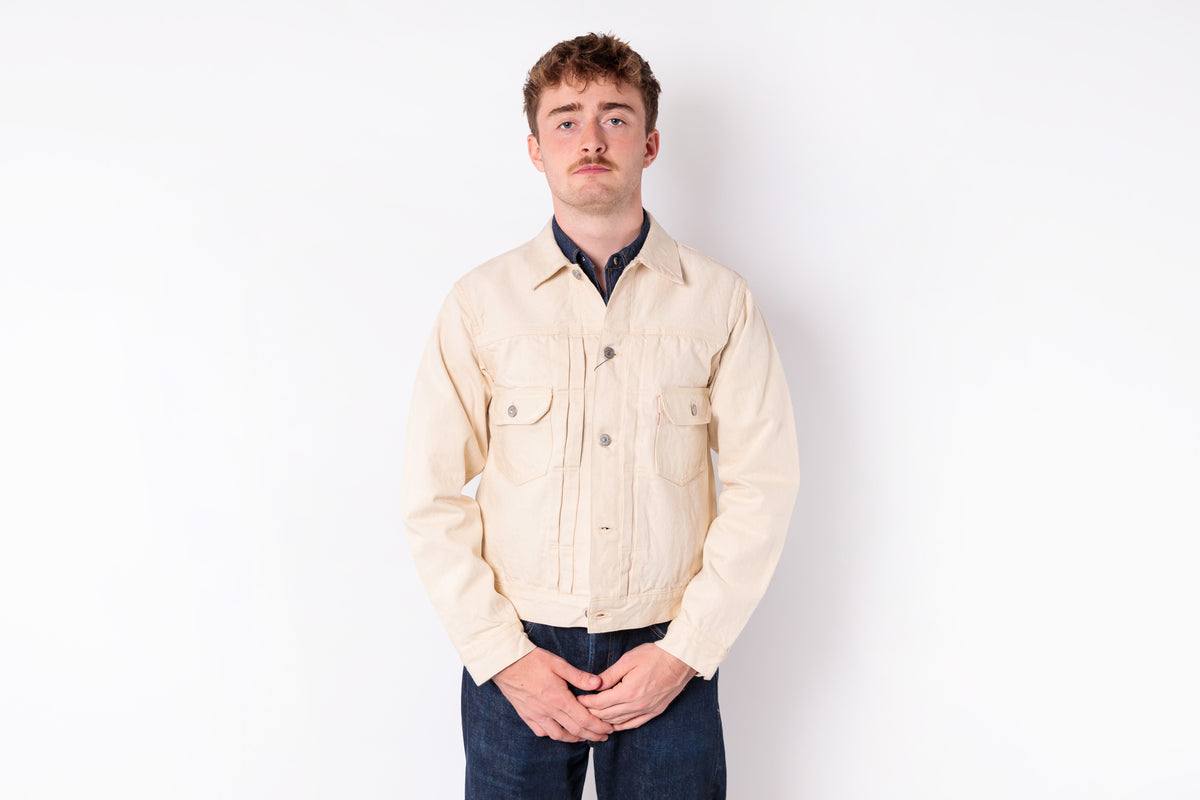 A man wearing a light beige canvas utility jacket over a dark navy collared shirt and dark blue jeans, standing against a plain white studio background.
