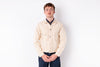 A man wearing a light beige canvas utility jacket over a dark navy collared shirt and dark blue jeans, standing against a plain white studio background.