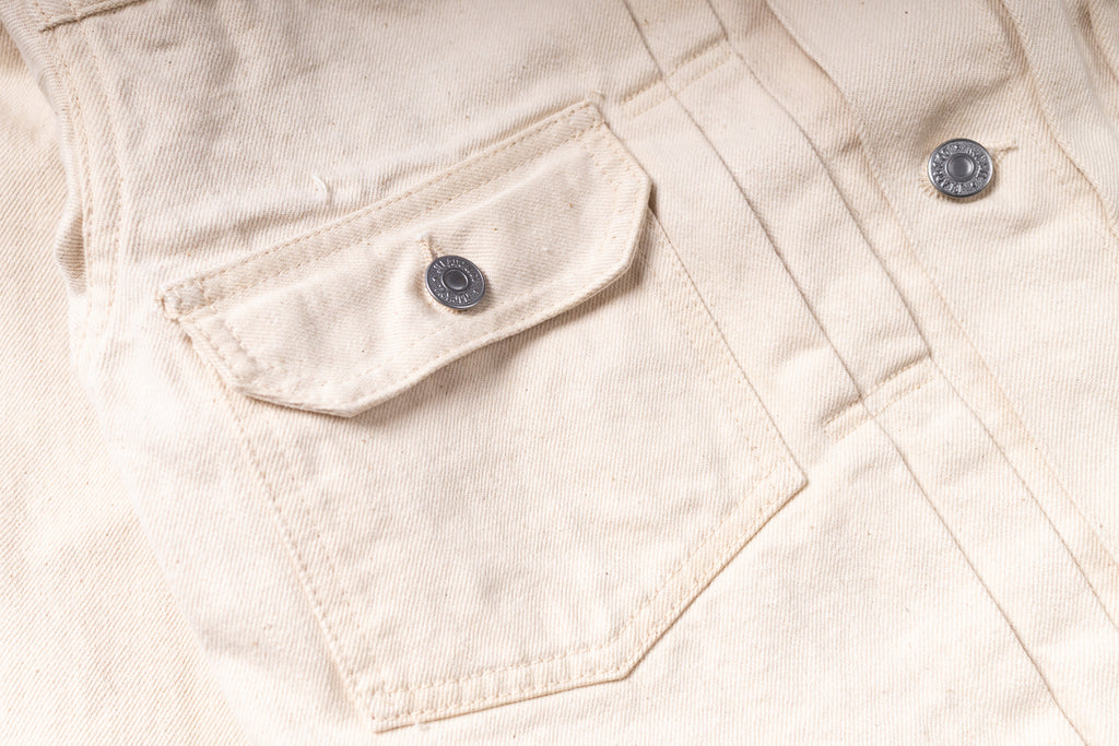 Close-up of a light beige canvas jacket featuring a chest pocket with a silver metal snap button and a matching snap button on the front placket.