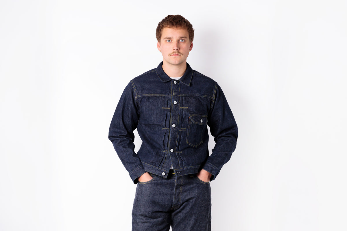 A man wearing a dark indigo denim jacket and matching dark jeans, standing against a plain white studio background with his hands in his pockets.